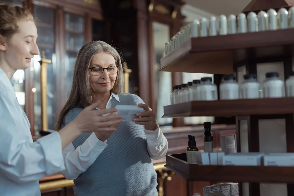A pharmacist showing a medicine box to an older customer beside the shelves at a pharmacy counter