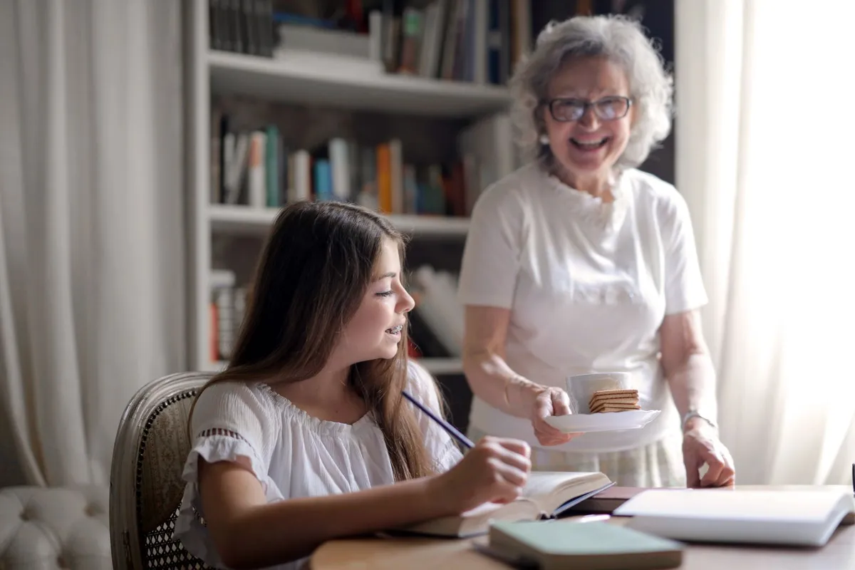 A smiling grandmother brings a cup and crackers to her granddaughter, who is doing homework at a table in front of a bookshelf