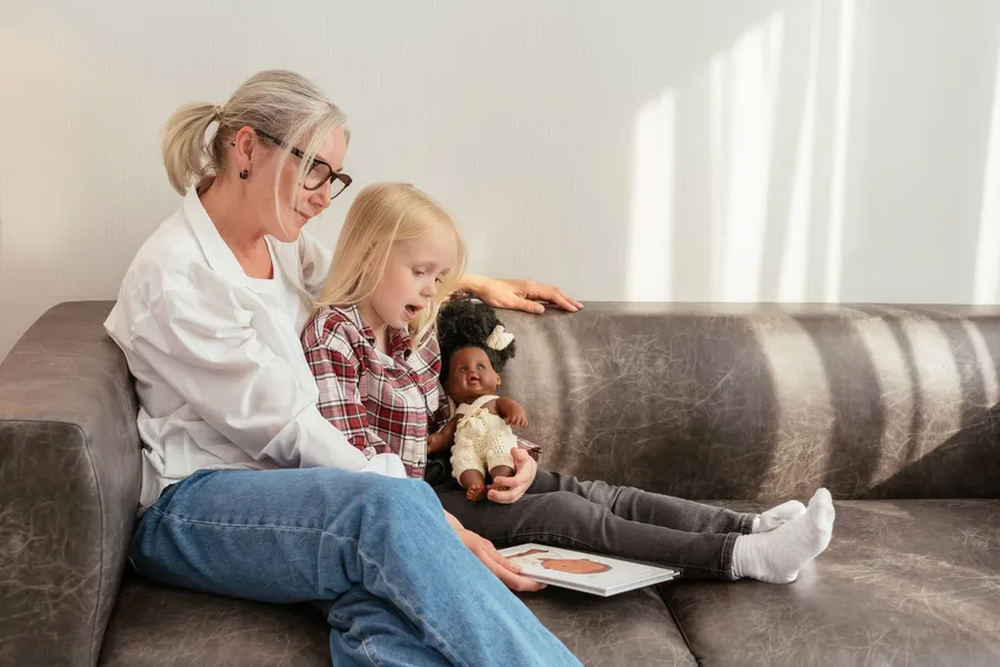 A grandmother with glasses sits on a leather couch reading a picture book with her young granddaughter, who holds a doll