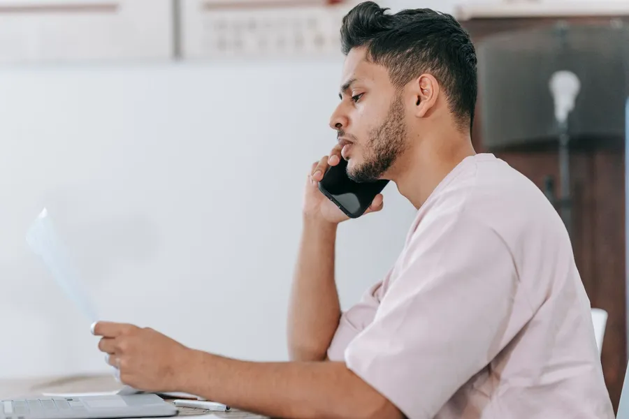 A man on a phone call reads a paper at a table with a laptop