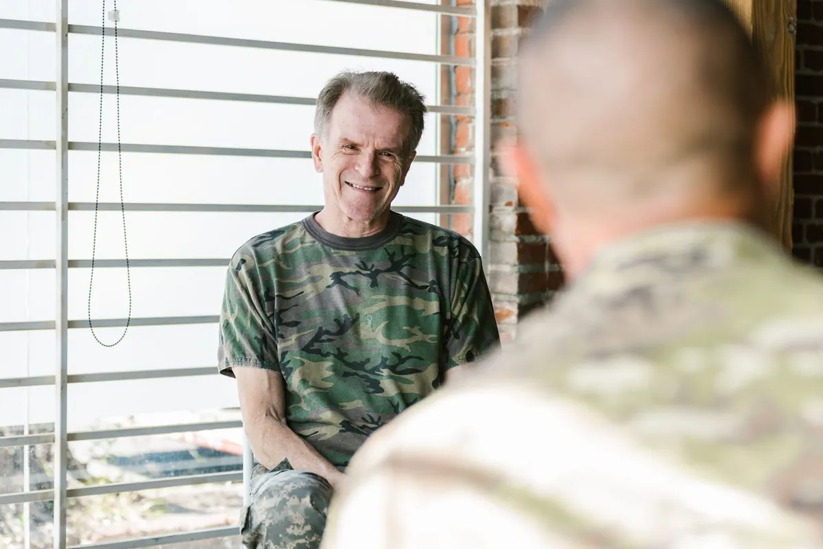 An older man in a camouflage T-shirt smiles while talking with a service member, seen from behind, by a bright window