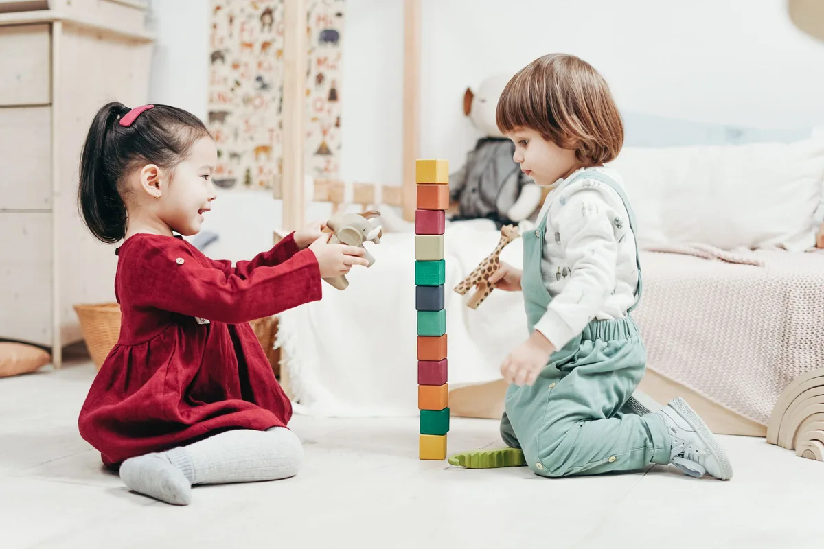 Two young children kneeling on a playroom floor, stacking colorful wooden blocks into a tower