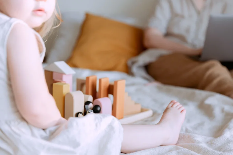 A toddler playing with wooden blocks on a bed while a parent works on a laptop in the background