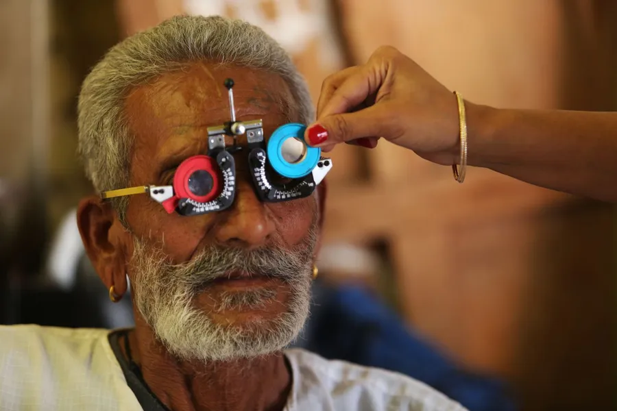 An older man looks through a trial frame with colored lenses while an eye care worker adjusts a lens during a vision test