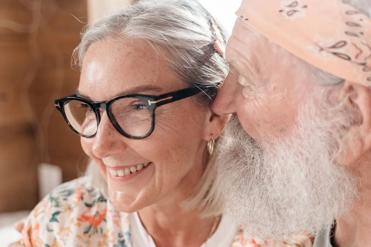 A smiling older woman in dark-framed eyeglasses leans close to an older man with a white beard