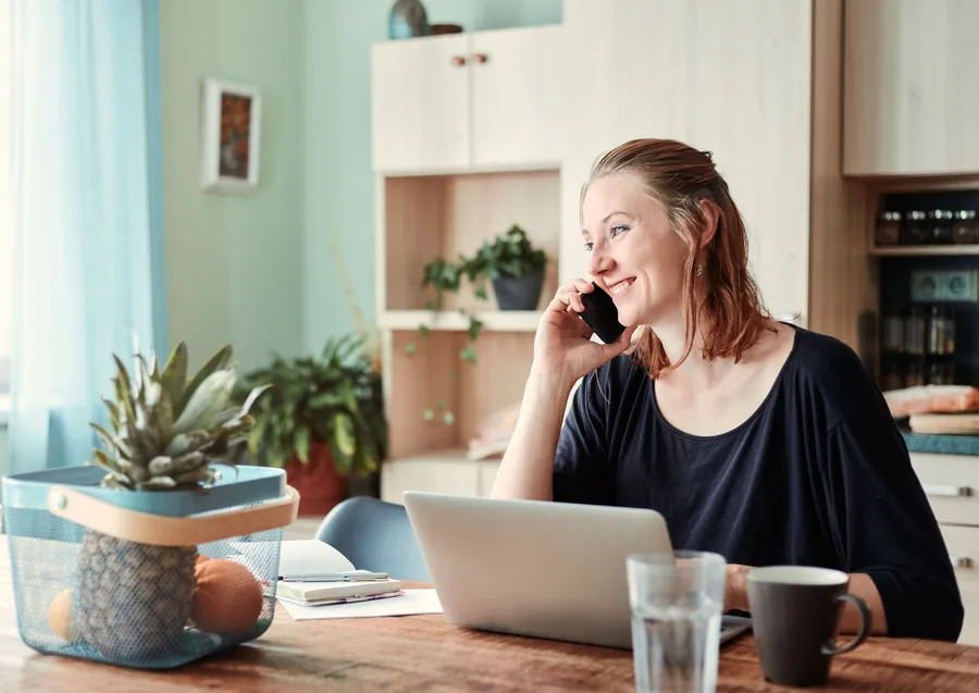 A woman on the phone at a kitchen table with a notebook, a laptop and a glass of water