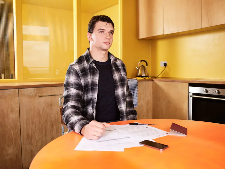A man sitting at a kitchen table with paperwork, a pen, a phone and an identity document