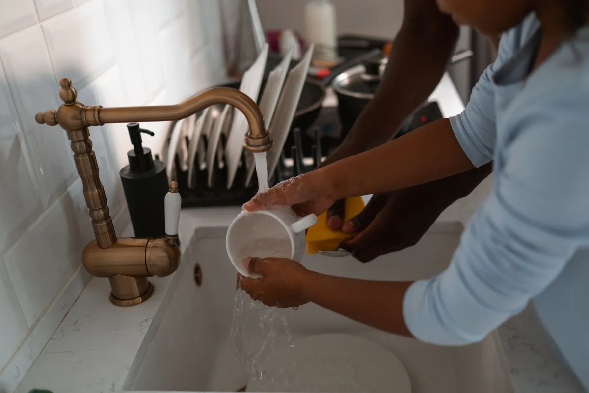 An adult and a child washing a mug together under running water at a kitchen sink