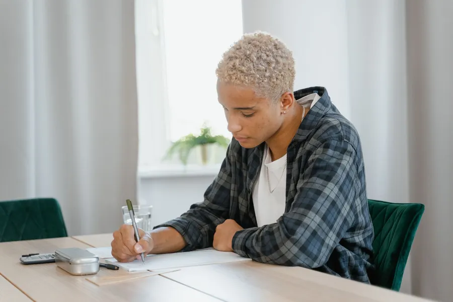 A young man in a plaid shirt writes on a notepad at a light wooden table, with a pencil case and a glass of water beside him