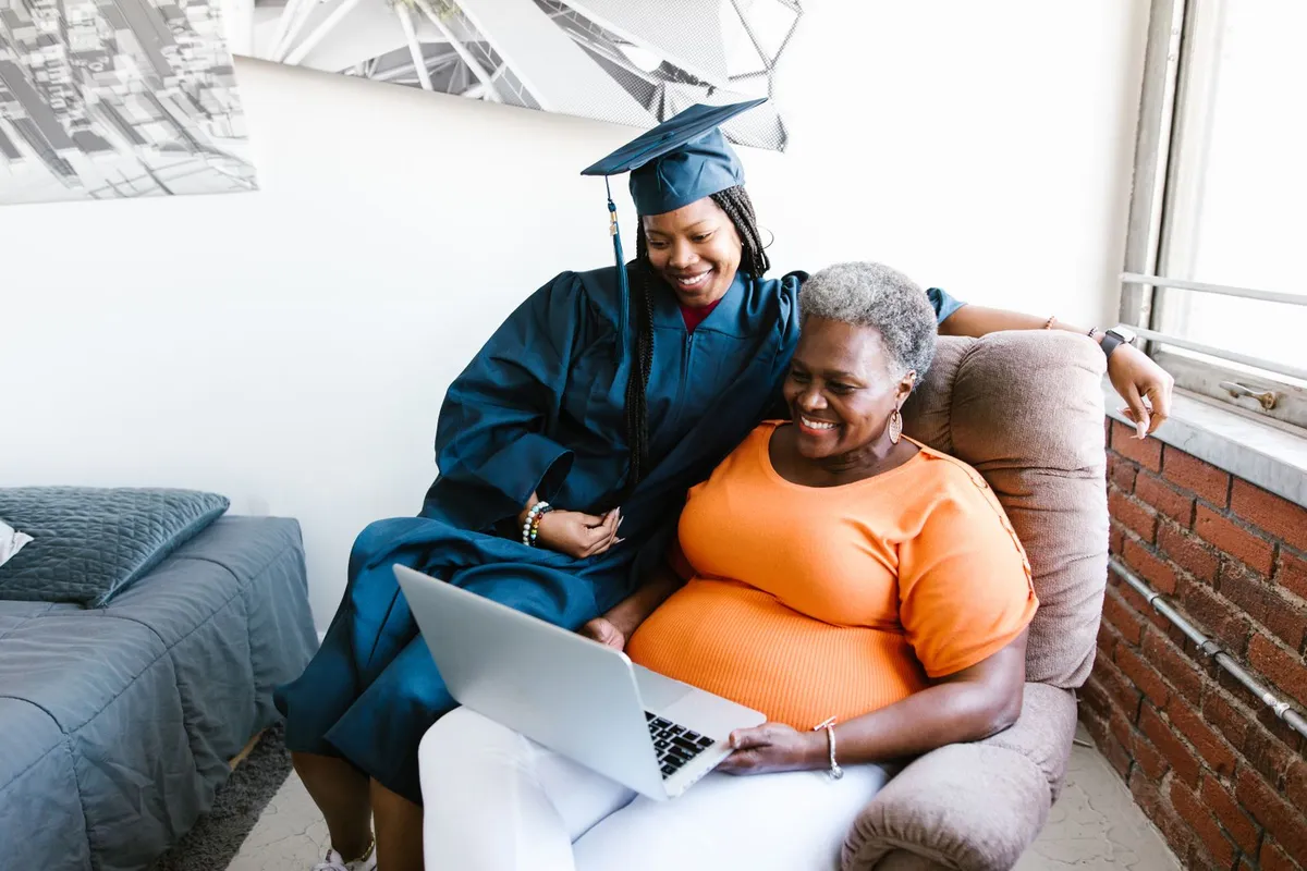 A smiling graduate in a cap and gown sits on the arm of a chair next to an older woman, and they look at a laptop together