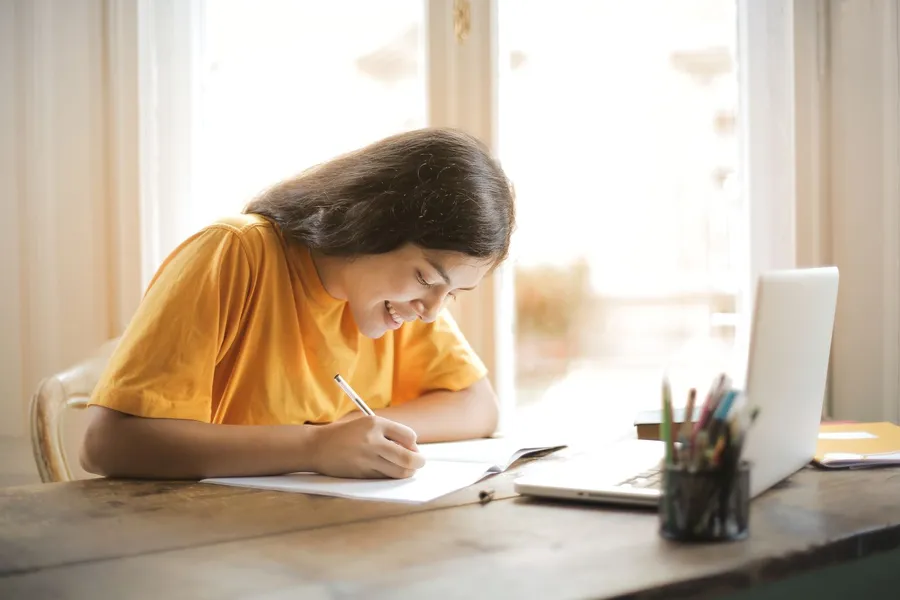 A smiling student in a yellow T-shirt writes in a notebook at a wooden table next to a laptop and a cup of pencils