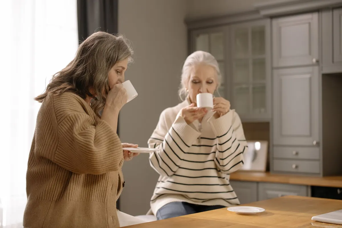 Two older women drink coffee together at a wooden kitchen counter in a bright home