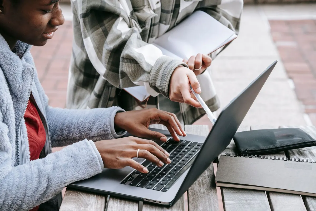 A smiling student in a fuzzy blue jacket types on a laptop at an outdoor table while a friend in a plaid shirt points to the screen with a pen