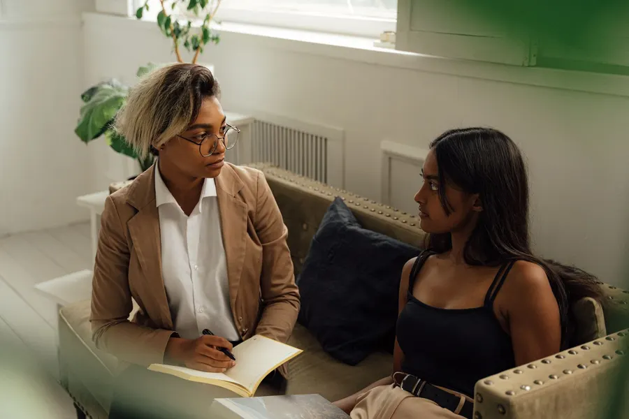 A woman in a tan blazer with a notebook talks with a young woman with long dark hair on a sofa in a bright room