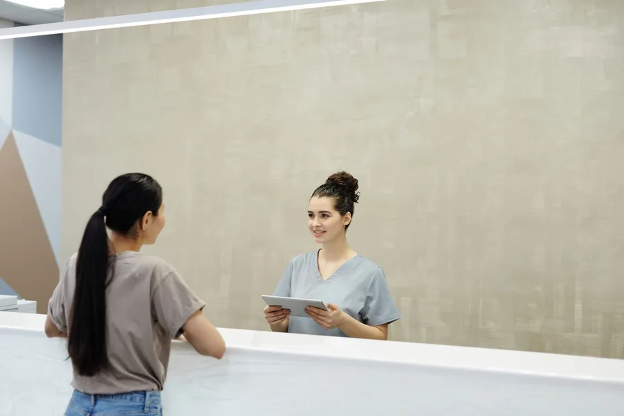 A patient standing at a hospital front desk, talking with a staff member holding a tablet