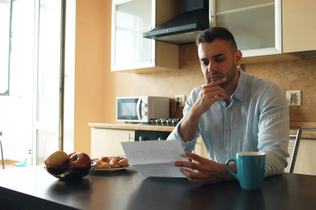 A man sitting alone at his kitchen table, reading a printed bill closely with a cup of coffee beside him