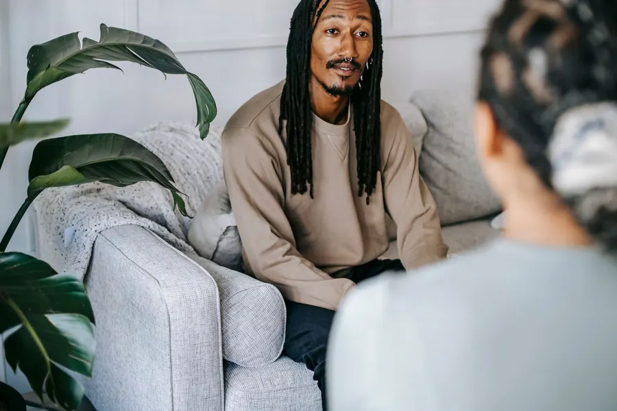 A man with long locs in a tan sweatshirt sits on a light gray sofa beside a large green plant, talking calmly with a person seen from behind
