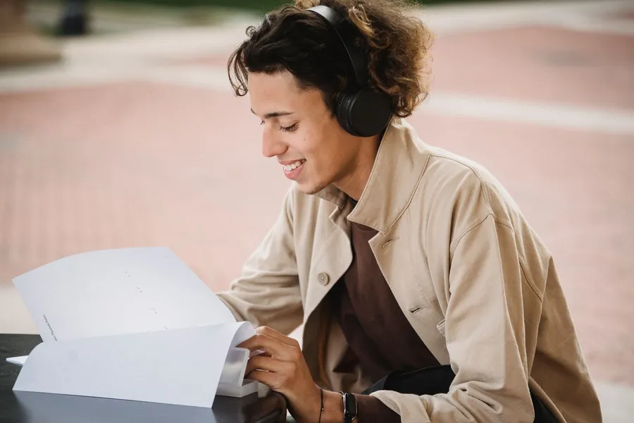 A smiling young man in headphones and a tan jacket reads through a stapled packet of papers at an outdoor table