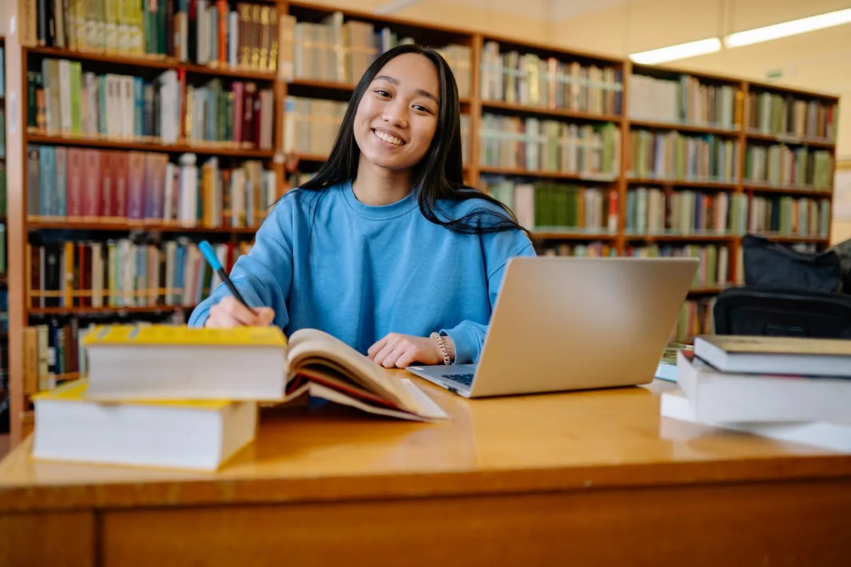 A smiling student in a blue sweatshirt sits at a library table with a laptop and a stack of books, holding a pen