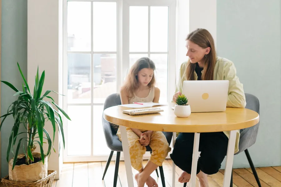 A parent and a child sitting at a sunlit table at home, working together at a laptop