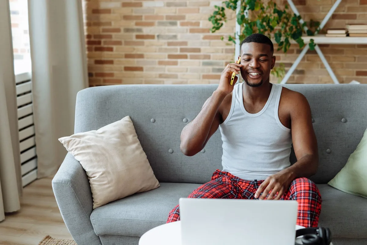 A man sitting on a sofa at home, talking on his mobile phone with a laptop open in front of him