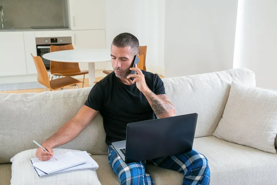 A man sits on a couch with a phone to his ear, writing notes in a notebook next to his laptop