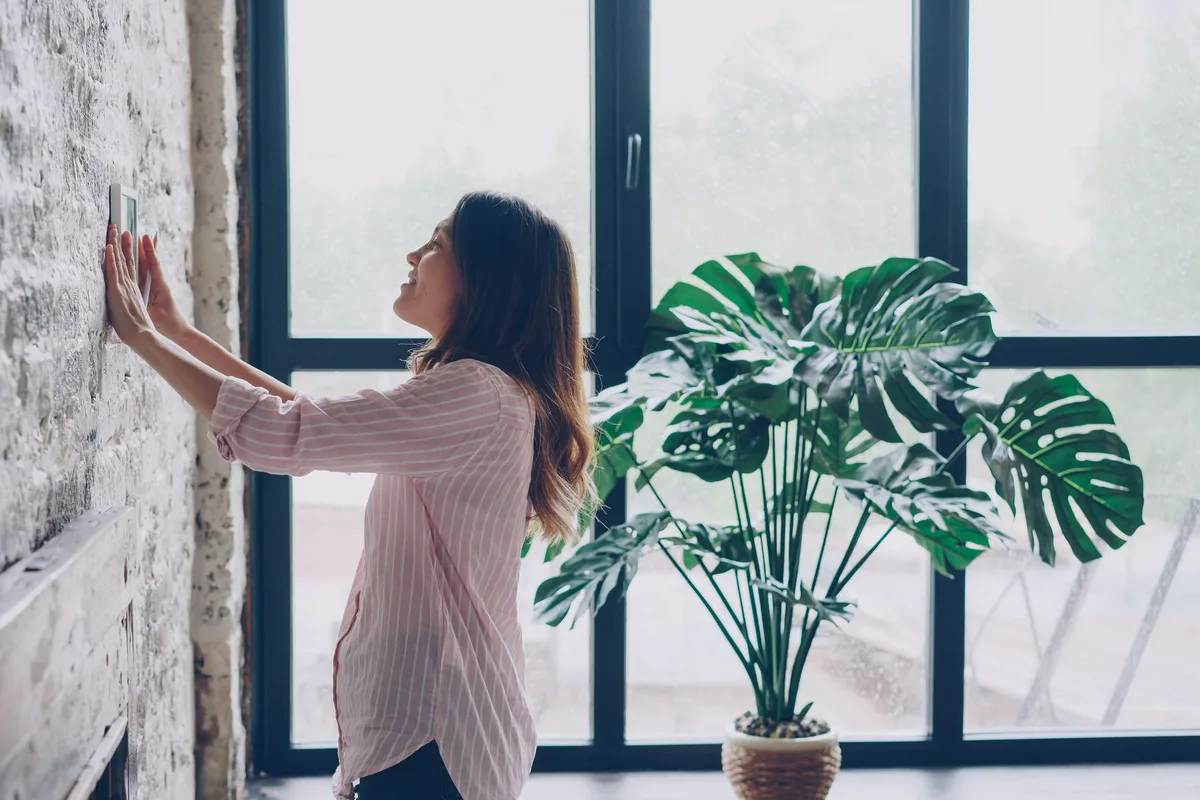 A woman reaching up to adjust the thermostat on the wall of a bright living room