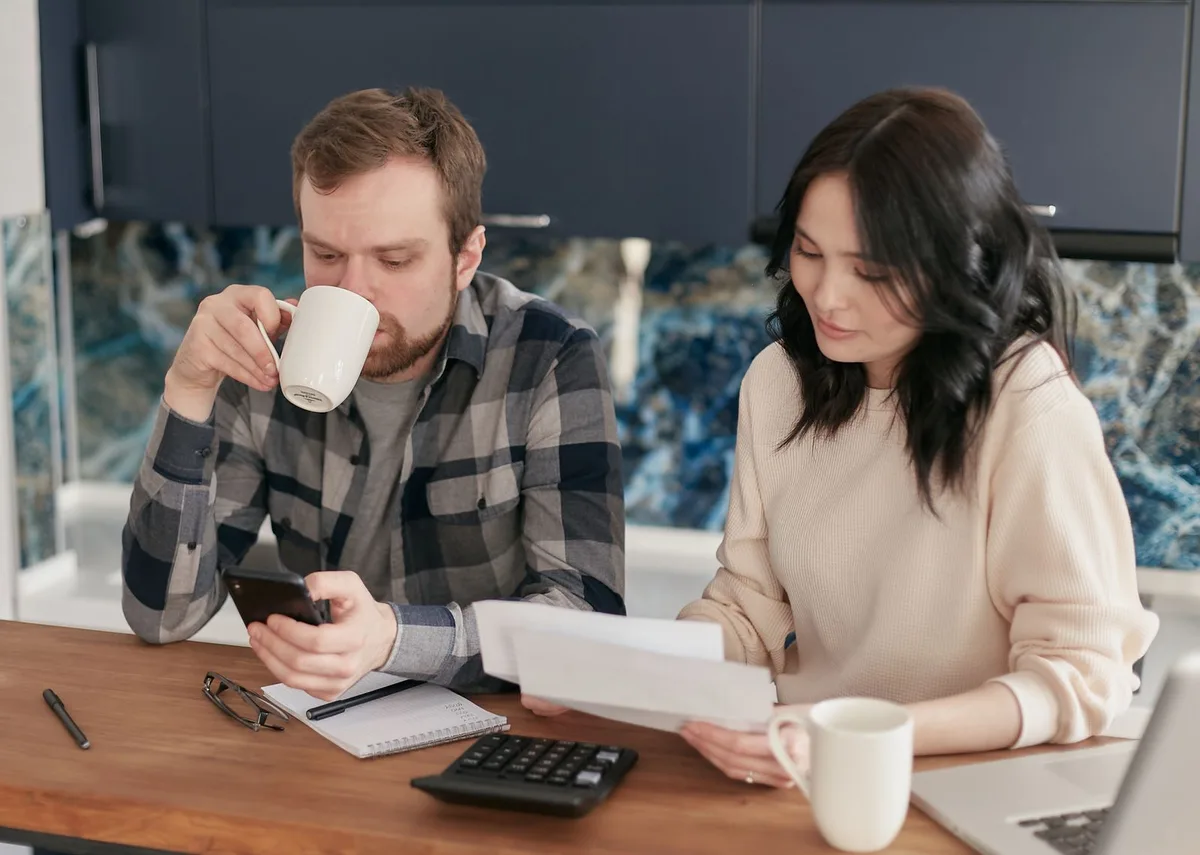 A couple at a kitchen table reviewing bills and paperwork with a calculator, notepad and phone