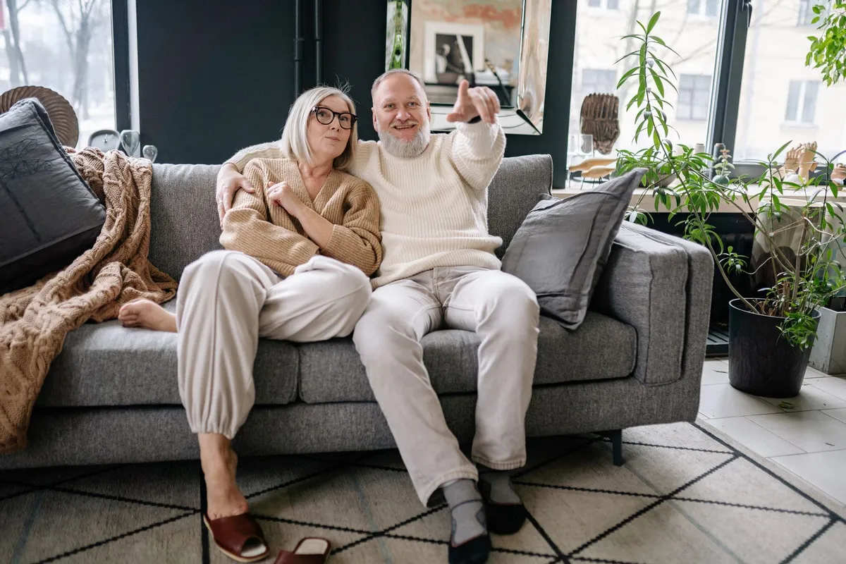 An older couple in knitted sweaters sit together on a gray couch at home, with bare winter trees visible through the windows behind them
