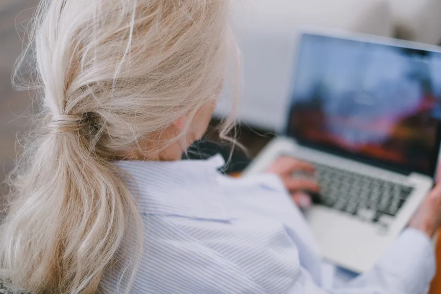 An older woman with gray hair, seen from behind, types on a laptop at home