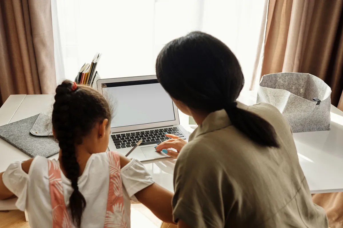 A mother and her young daughter sit side by side at a desk by the window, looking at a laptop together