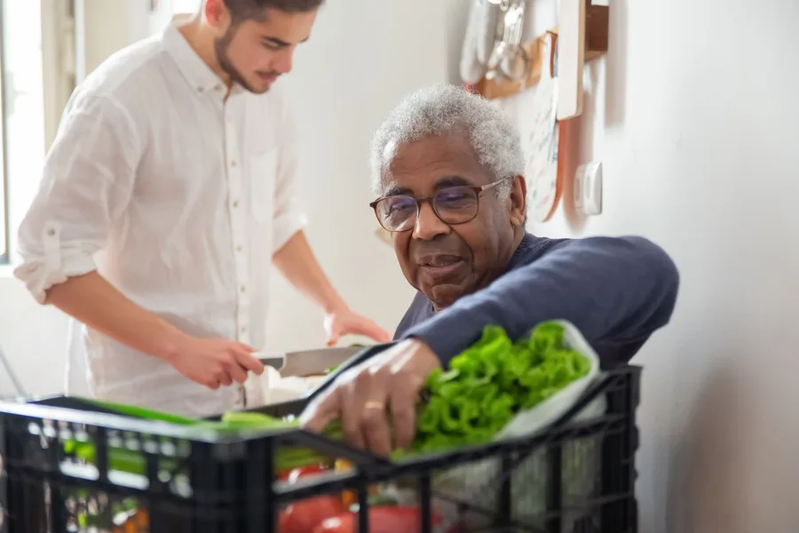 An older man unpacking a crate of fresh greens on his kitchen counter while a younger helper stands beside him
