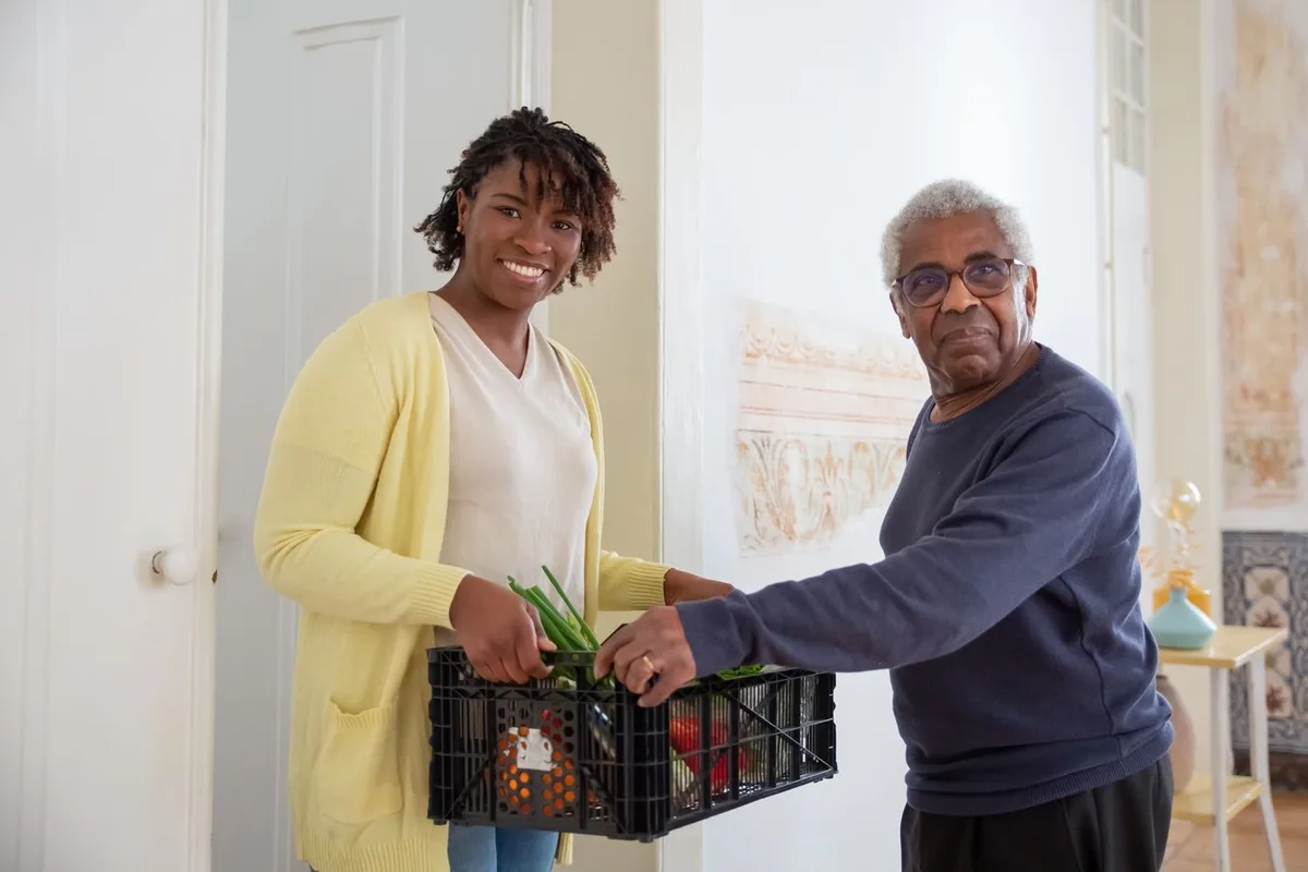 A volunteer handing a crate of fresh vegetables to an older man at the door of his home
