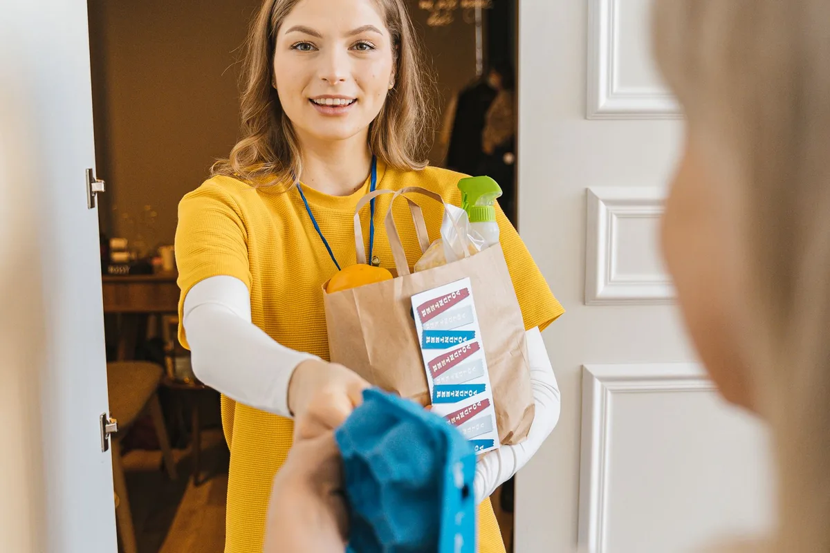 A volunteer in a yellow shirt handing a paper bag of groceries to an older person at the door of their home