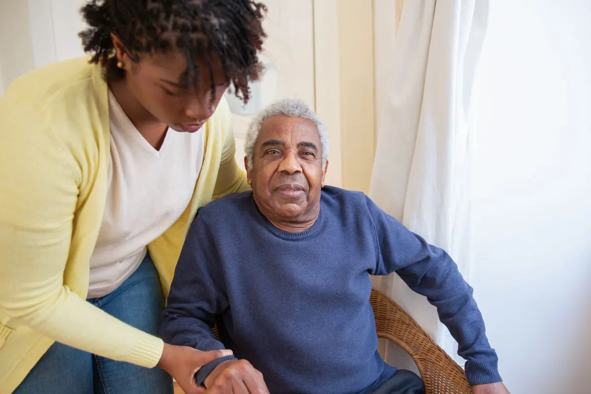 A younger woman in a yellow cardigan helps an older man settle into a wicker chair at home; he looks toward the camera with a calm expression