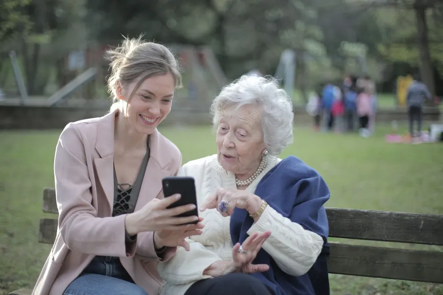 A young woman and an older woman sit on a park bench looking at a phone together and smiling