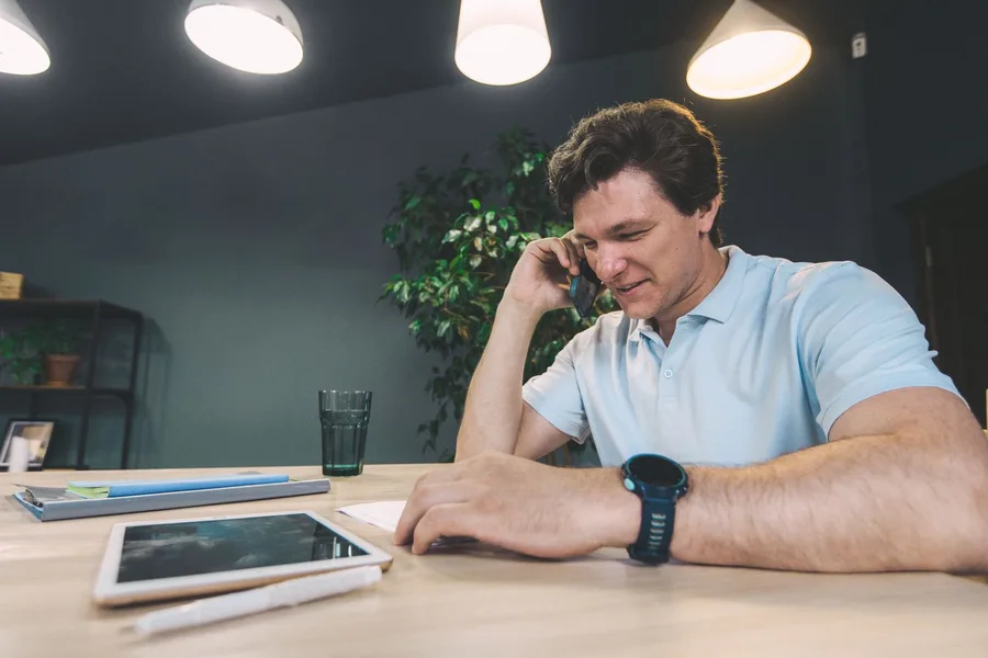 A man talks on the phone at a table with a tablet and a notepad in front of him