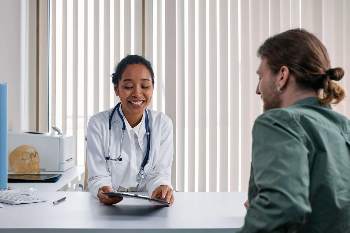 A doctor in a white coat smiling while going over notes on a tablet with a patient across the desk