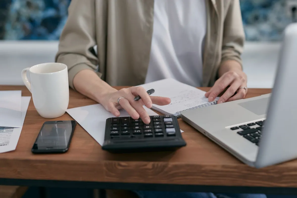 A person at a wooden desk working through numbers with a calculator, a notebook, and a laptop