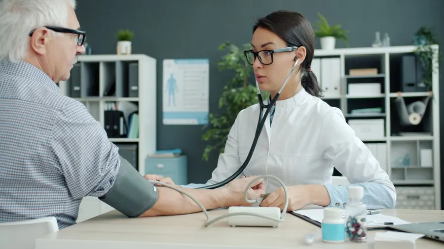 An older man in a checked shirt has his blood pressure taken by a doctor at a desk in a bright clinic
