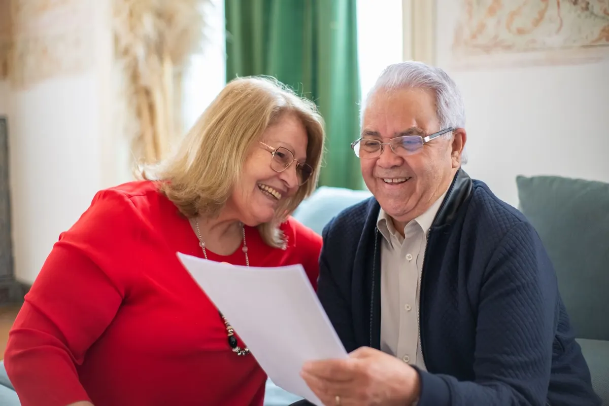 An older couple sitting on a couch, smiling as they read a printed letter together