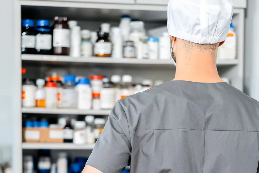 Shelves of medicine bottles behind a pharmacy worker in gray scrubs