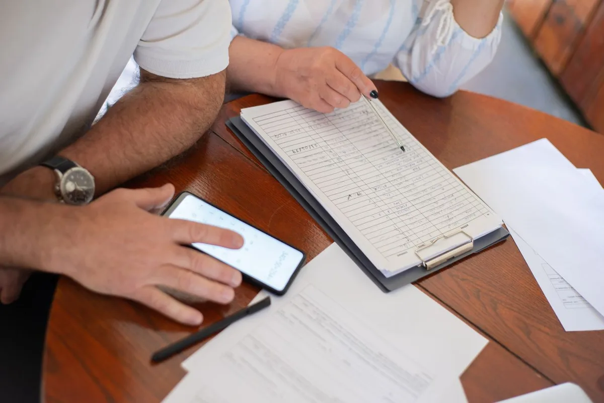 Two people at a kitchen table going over a printed application form on a clipboard, with a phone calculator open beside it
