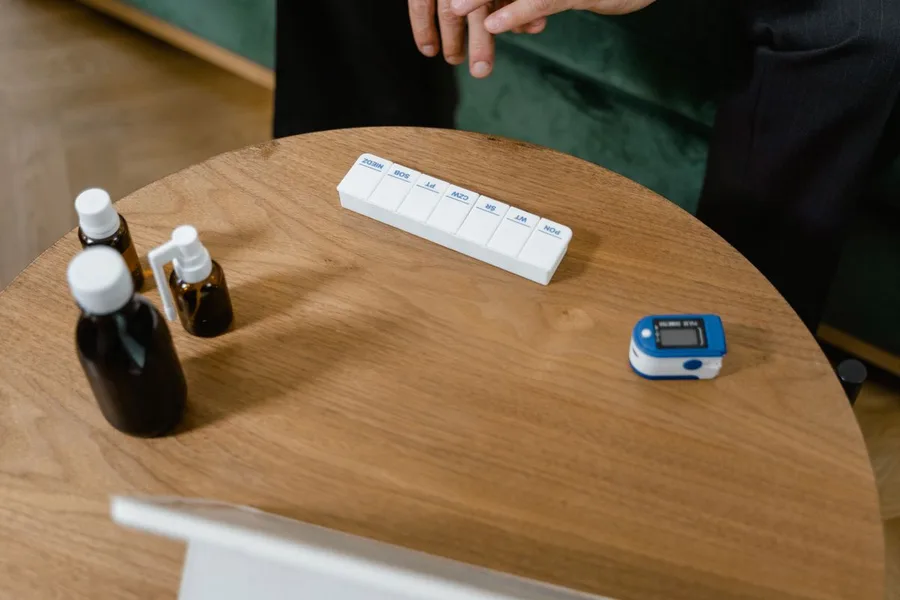 A weekly pill organizer, two medicine bottles and a fingertip pulse oximeter on a wooden side table