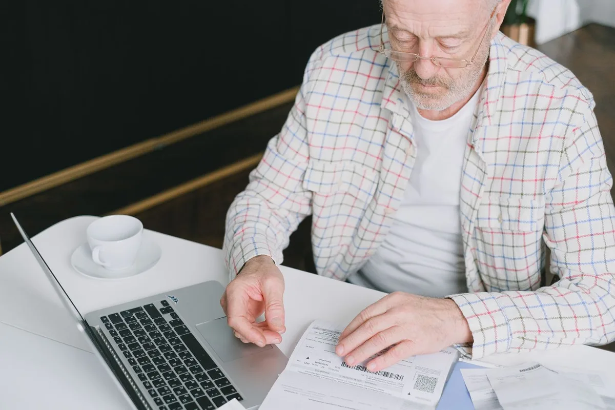 An older man at a kitchen table reading paperwork next to a laptop and a cup of coffee