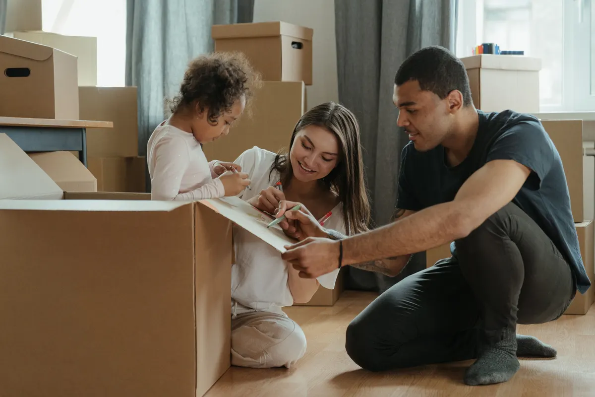 A young family kneels among moving boxes in a new home, smiling as they write on a sheet of paper together