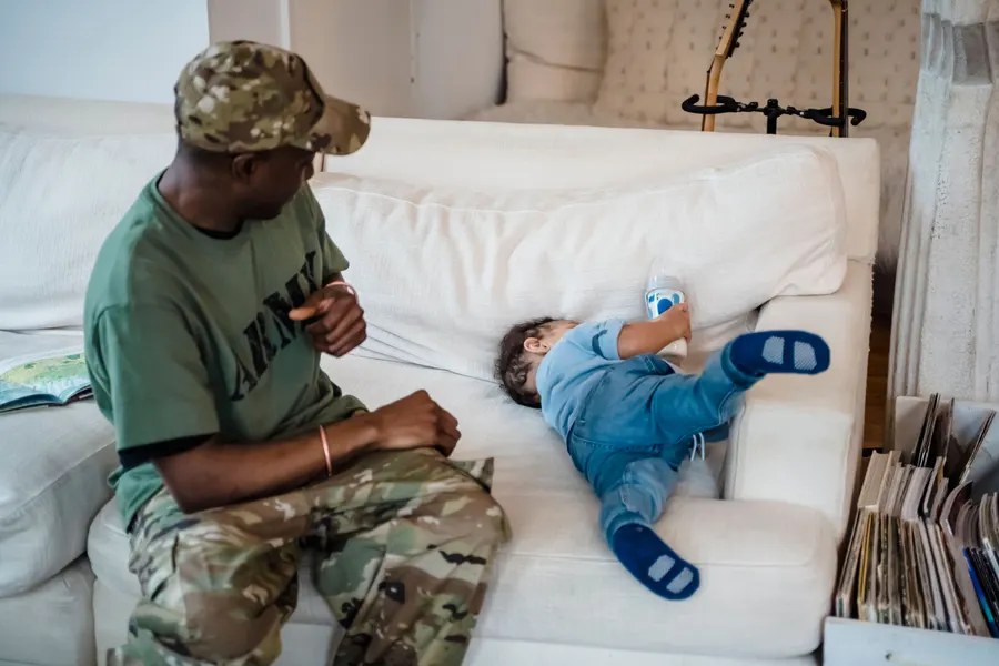 A service member in a camouflage cap and an Army T-shirt sits on a white sofa and watches a toddler holding a bottle