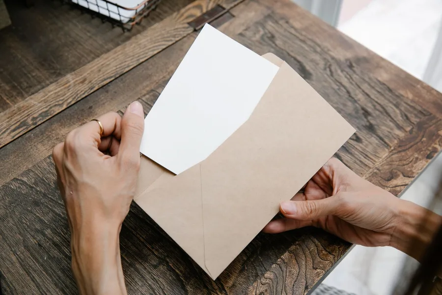 Two hands drawing a plain sheet of paper out of a kraft paper envelope on a wooden table