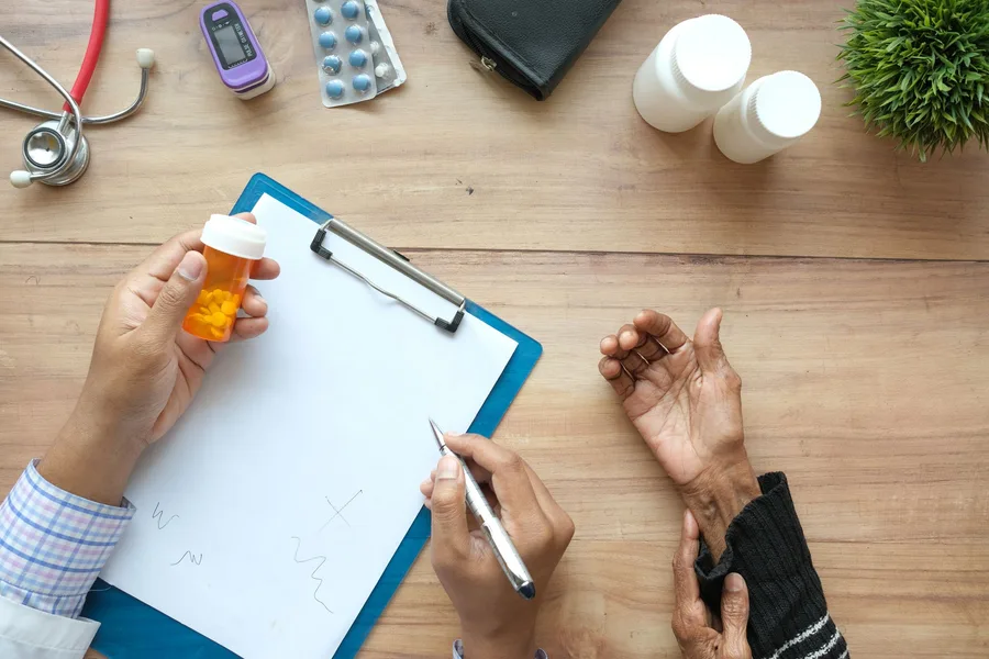 A clipboard, an orange prescription vial, pill bottles and a stethoscope on a wooden desk as a clinician talks with an older patient