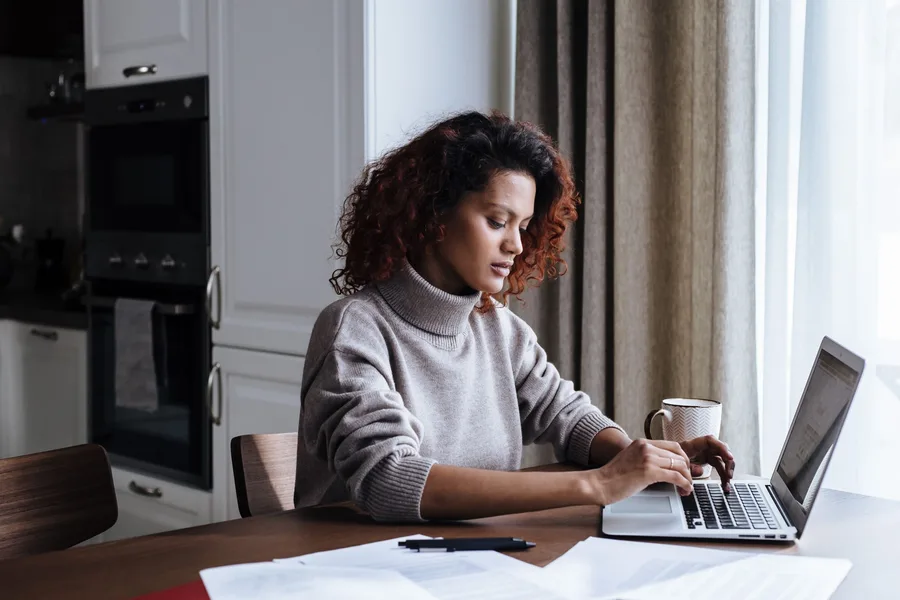 A woman at a kitchen table reading printed documents beside an open laptop